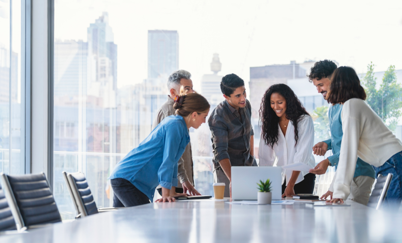 Team of business people working together in a board room. They are all standing and talking. There is technology on the table including a laptop computer. Ethnically diverse group. There is also some paperwork.